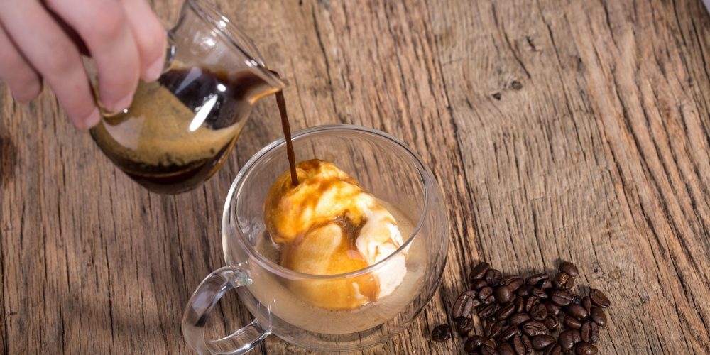 Affogato, pouring espresso coffee to Vanilla ice cream in double walled glass italian dessert, with coffee beans on the rustic wooden table